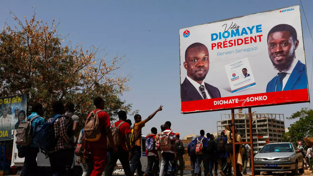 Students walks as they pass an electoral billboard of the Senegalese presidential candidate Bassirou Diomaye Faye, who is backed in the March 24 election by opposition leader Ousmane Sonko, in Dakar, March 20, 2024